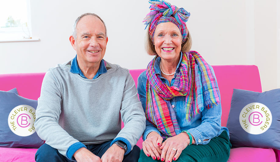 A late middle-aged white couple sat on a bright pink sofa. She is wearing a brightly coloured scarf, blue shirt and green trousers while he is dressed in a light grey jumper over blue shirt and trousers.