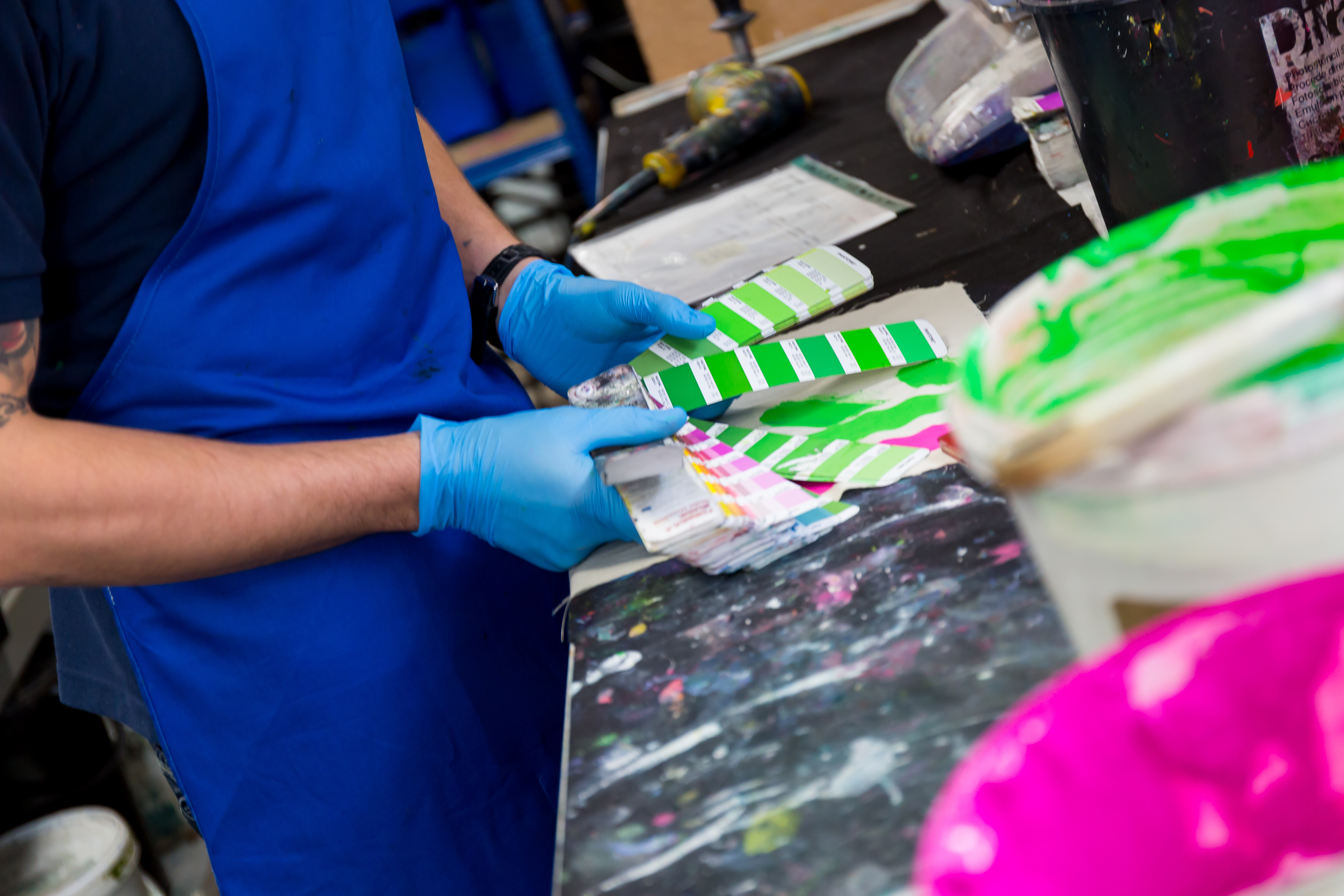 Buckets of colours prepared for screen print wait on a bench while a person checks them against a Pantone reference book.