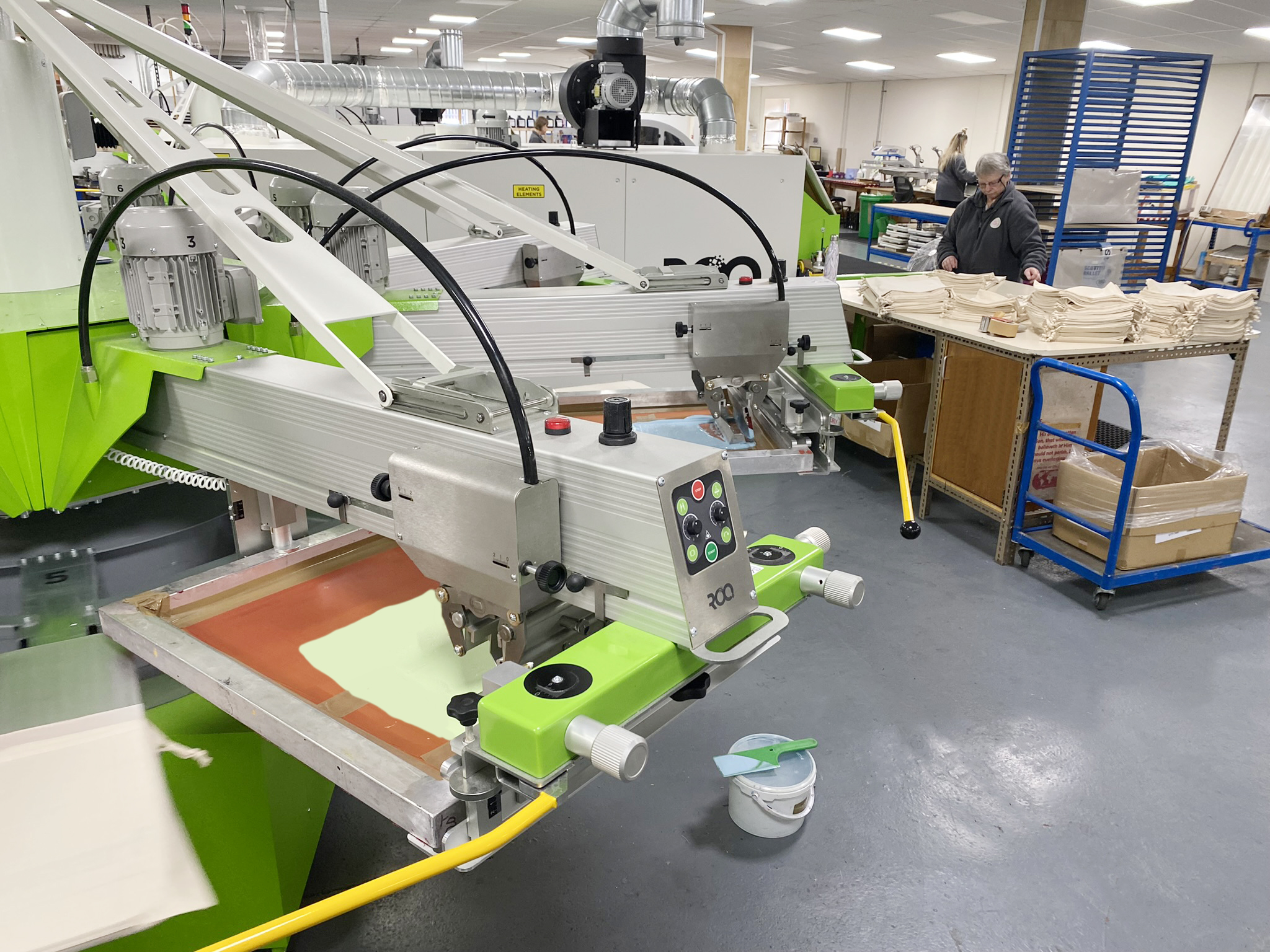 Carousel machine for automated screen printing, in a clean and tidy workshop. Behind the machine a person stands checking printed cotton bags at a bench and stacking them neatly ready for packing. In the background of the photo other people are working at different print process machines.