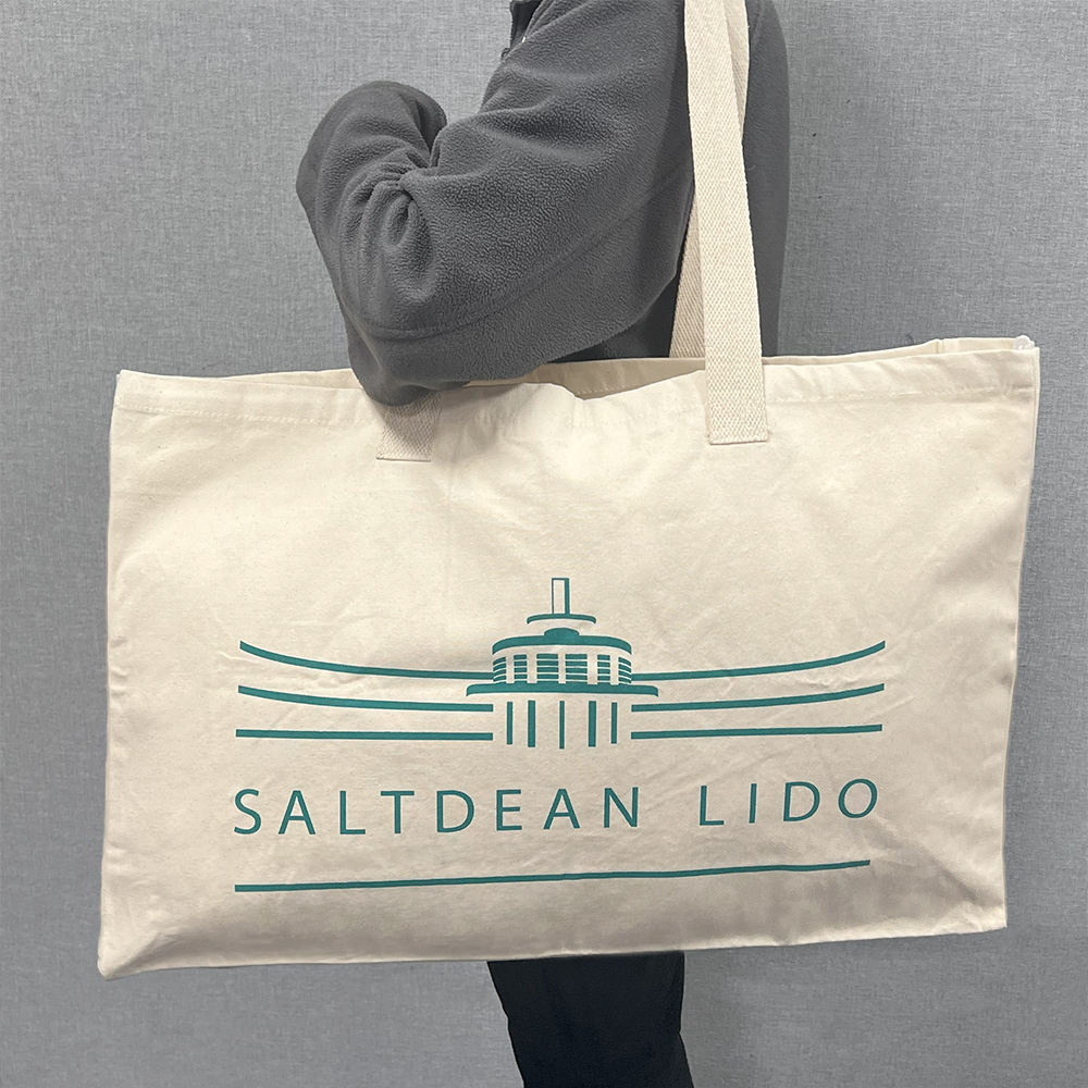 Large beige cotton canvas bag, printed with the logo of Saltdean Lido, which covers most of the width of the bag. The bag is stuffed full and being carried over a person's shoulder.