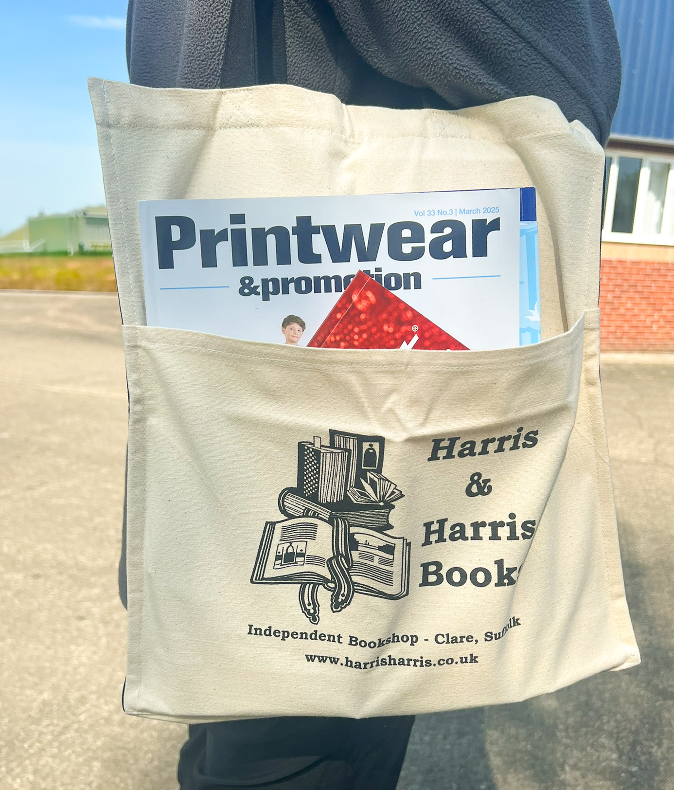 Cotton canvas tote bag carried over a person's shoulder. The bag has a full width front pocket printed with the name and details of a bookshop and is being used to hold a couple of magazines.