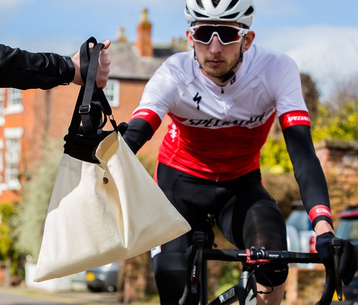 A cyclist in branded racing gear is approaching the camera. Ahead of him, an arm is extended into the shot, holding out a beige canvas bag for the cyclist to take on his way past.