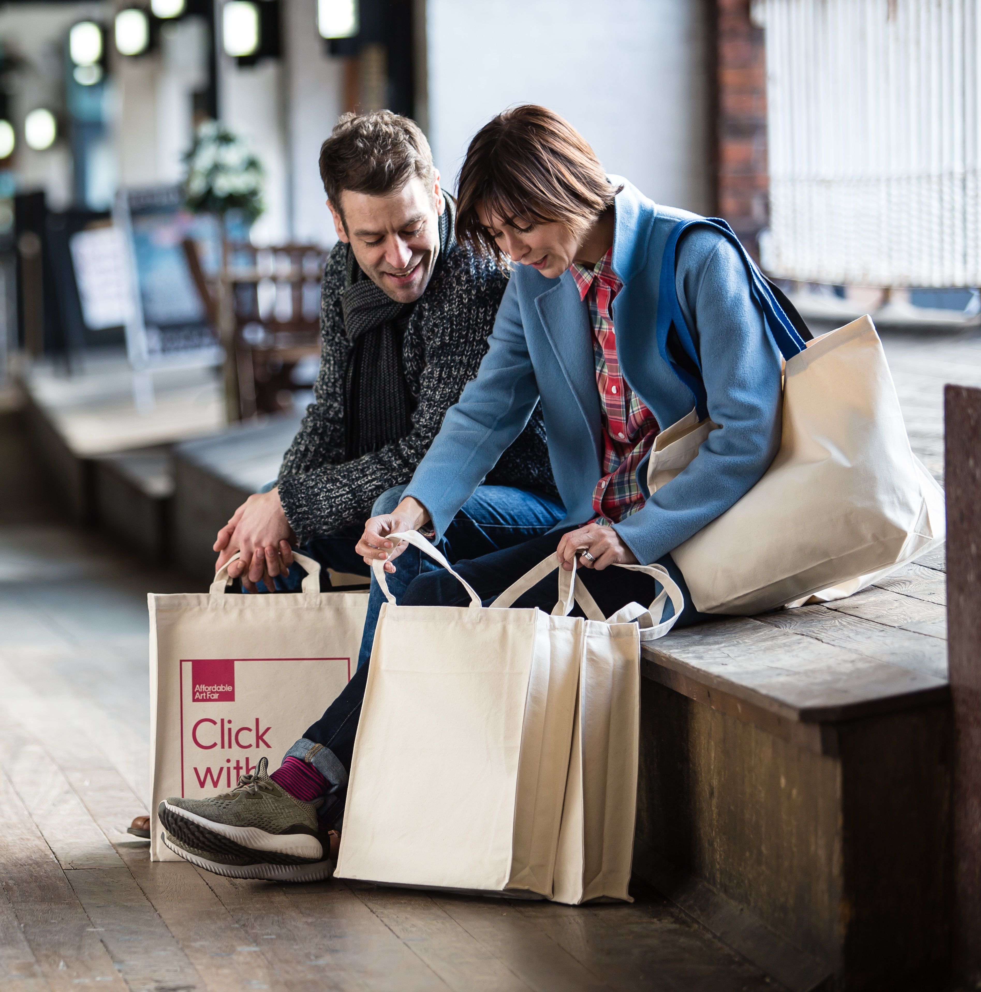A man and woman sat down outdoors, surrounded by canvas tote bags. They are both looking into one of the bags which the woman is holding, and smiling or laughing as they do so.