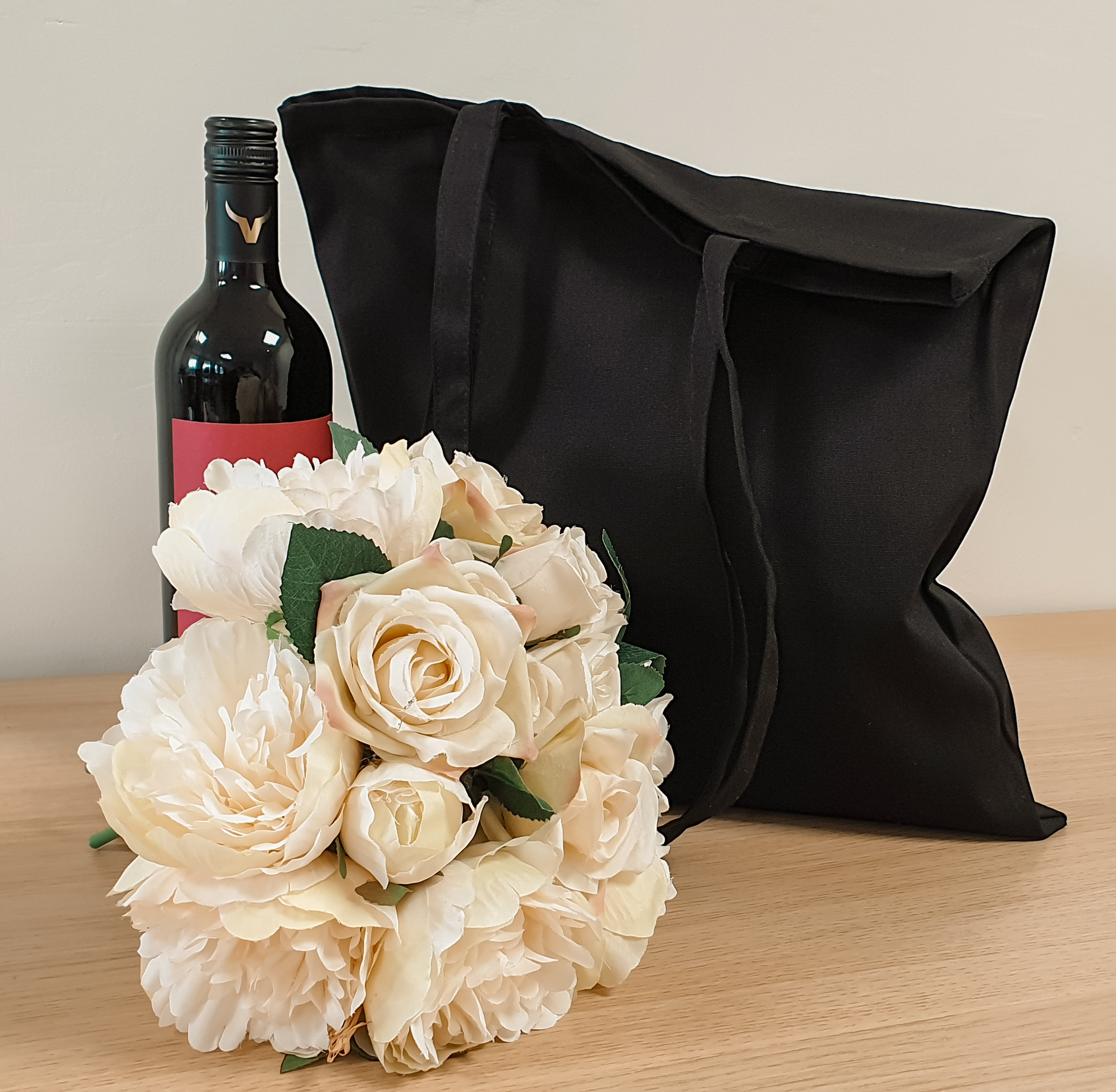 A black tote bag sits on a light coloured table with a bottle of wine next to the bag and a large bunch of creamy white flowers in front.