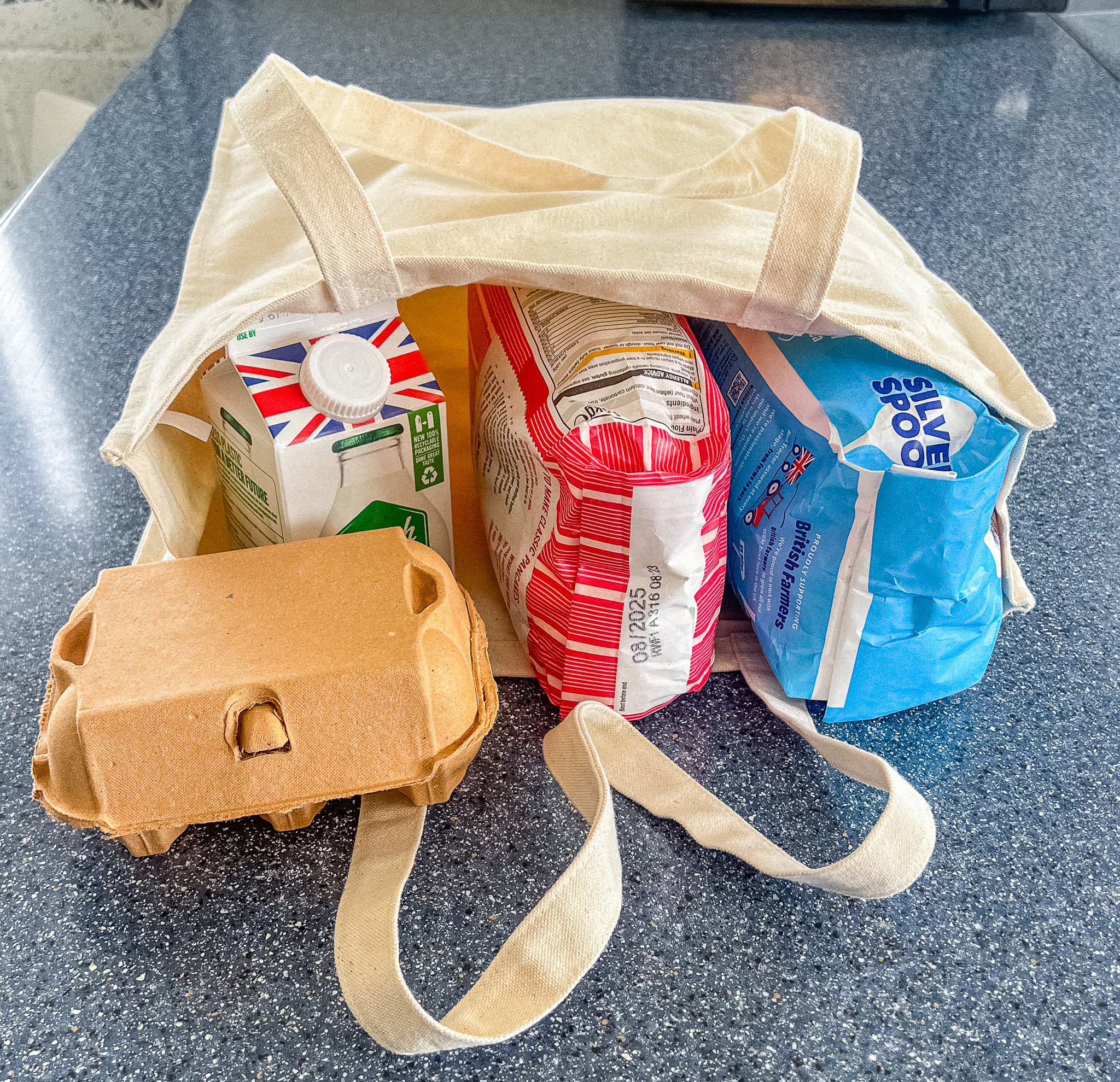 Beige cotton canvas tote bag lying on its side on a kitchen worktop with baking ingredients such as bags of flour and sugar inside.