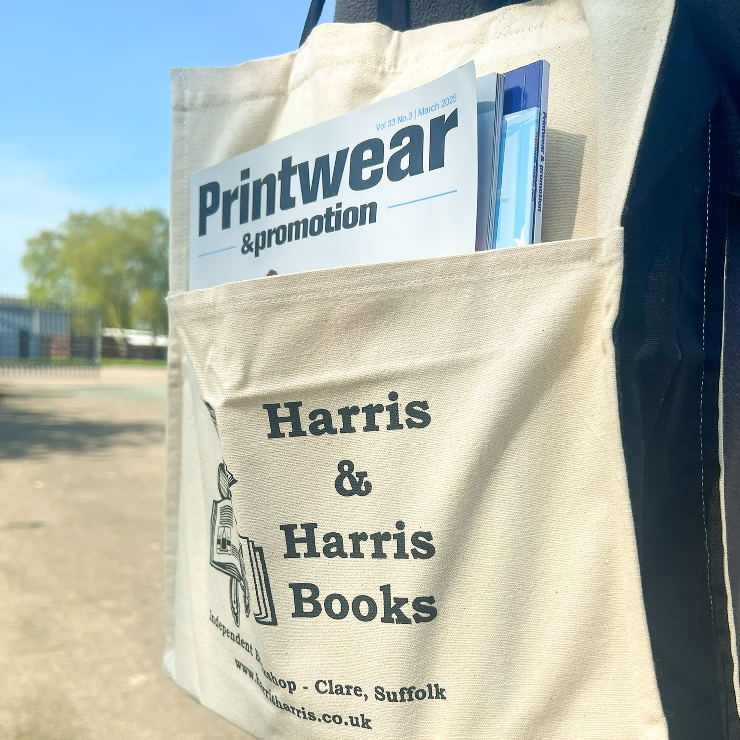 Tote bag carried over shoulder. Front pocket printed with name & details of a bookshop and holds magazines.