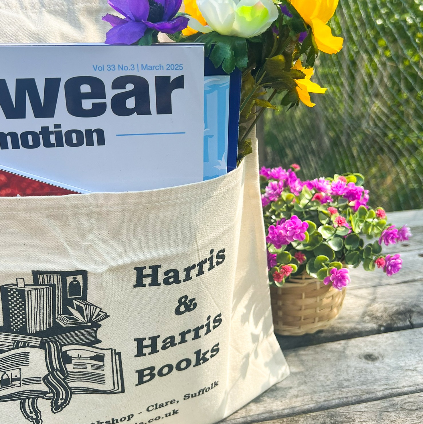 Tote bag sat on wooden table next to flowers. Front pocket printed with name & details of a bookshop and holds magazines.