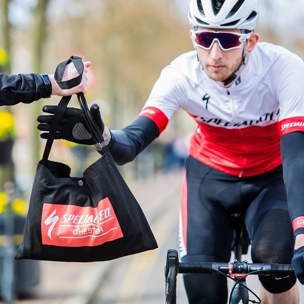 A cyclist in branded clothing approaches the camera, one hand extended to take a branded black canvas bag which is held out to them by someone standing just out of shot.