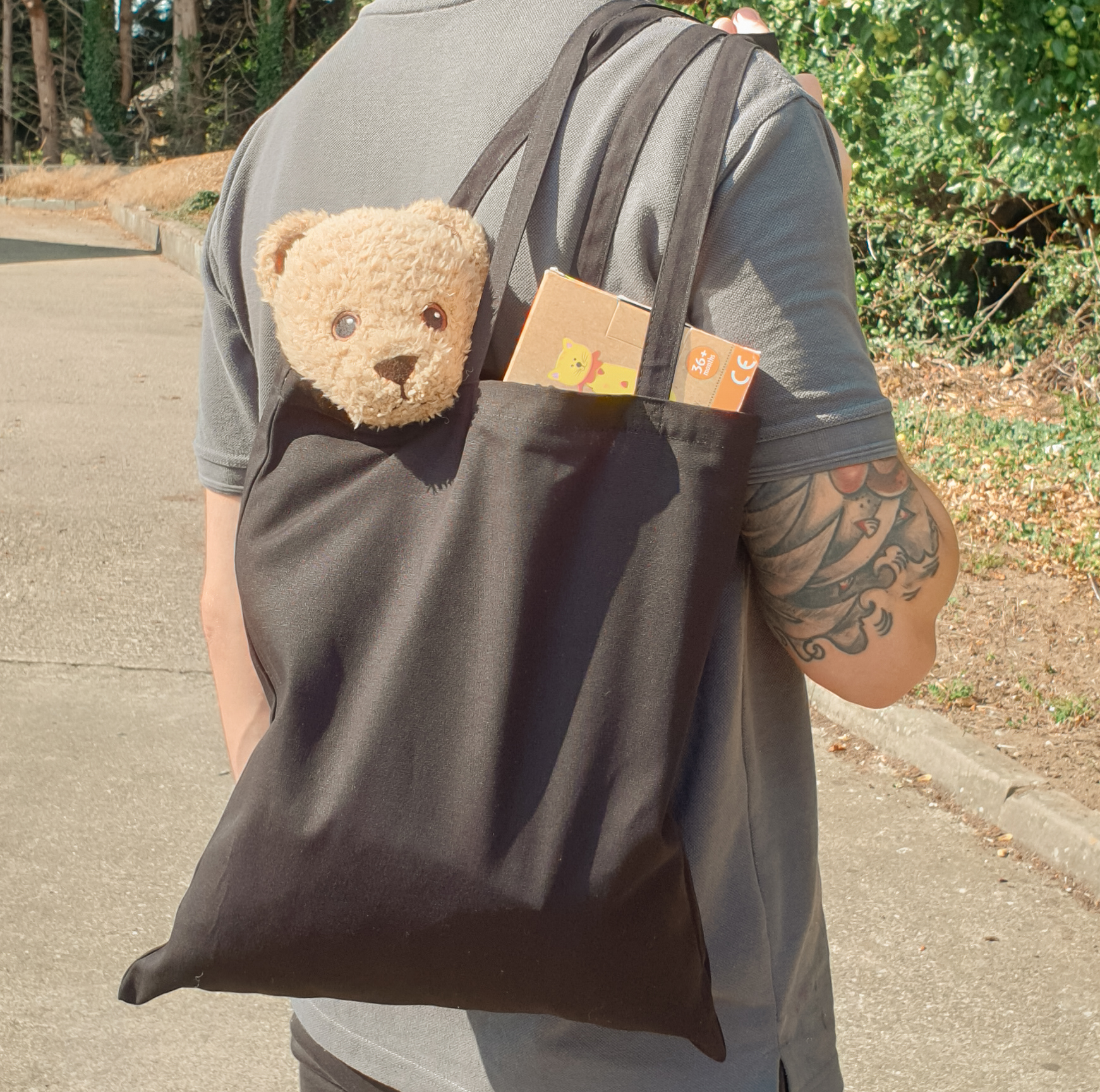 A man in a grey polo shirt holds a black canvas tote bag over his shoulder. Sticking out of the top of the bag are children's toys: something in a tastefully colourful box, and a teddy bear.