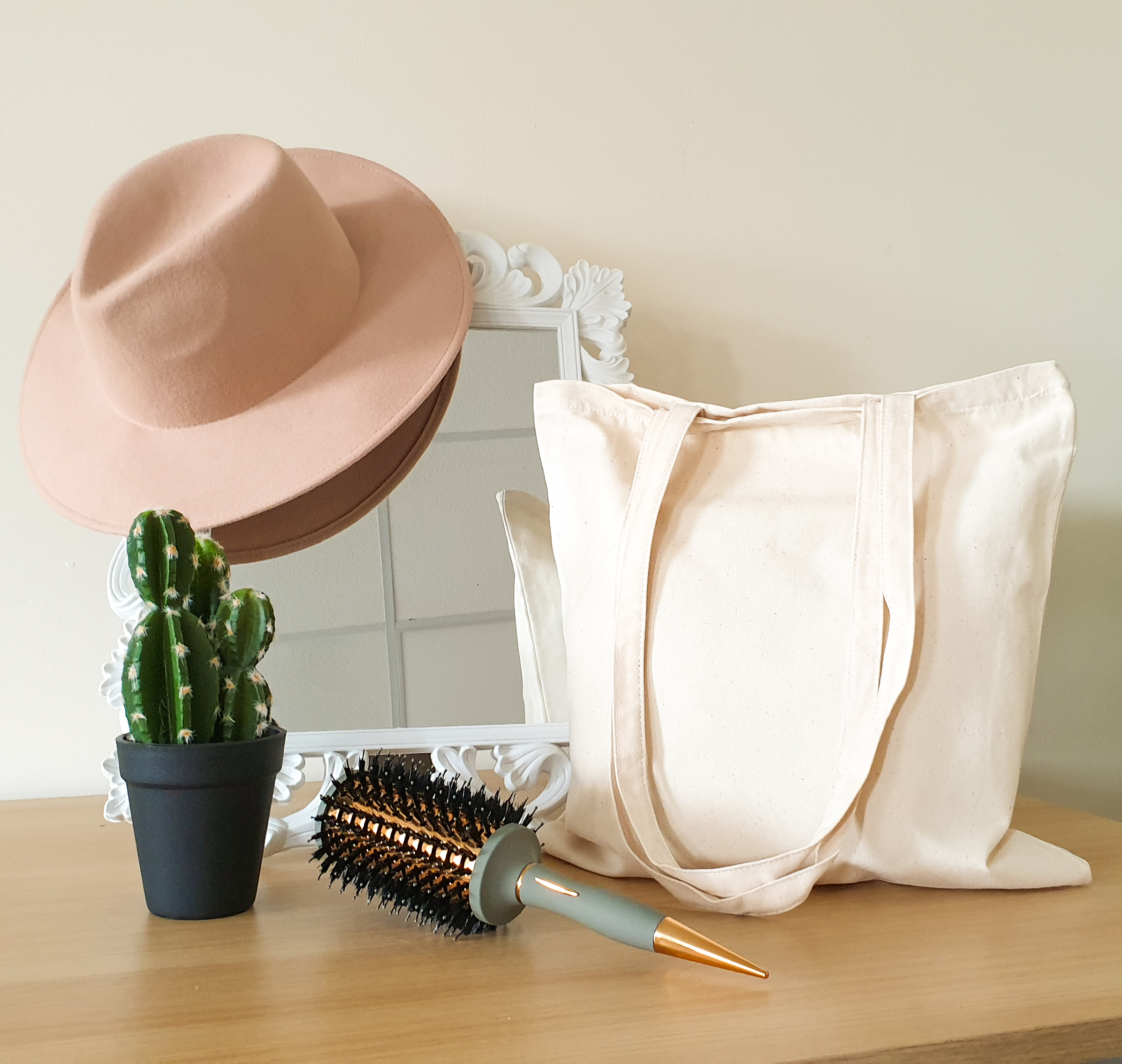 A beige tote bag sits by a white-framed mirror. A felt fedora style hat hangs over one corner of the mirror with a hair brush in front and a small cactus plant to one side.