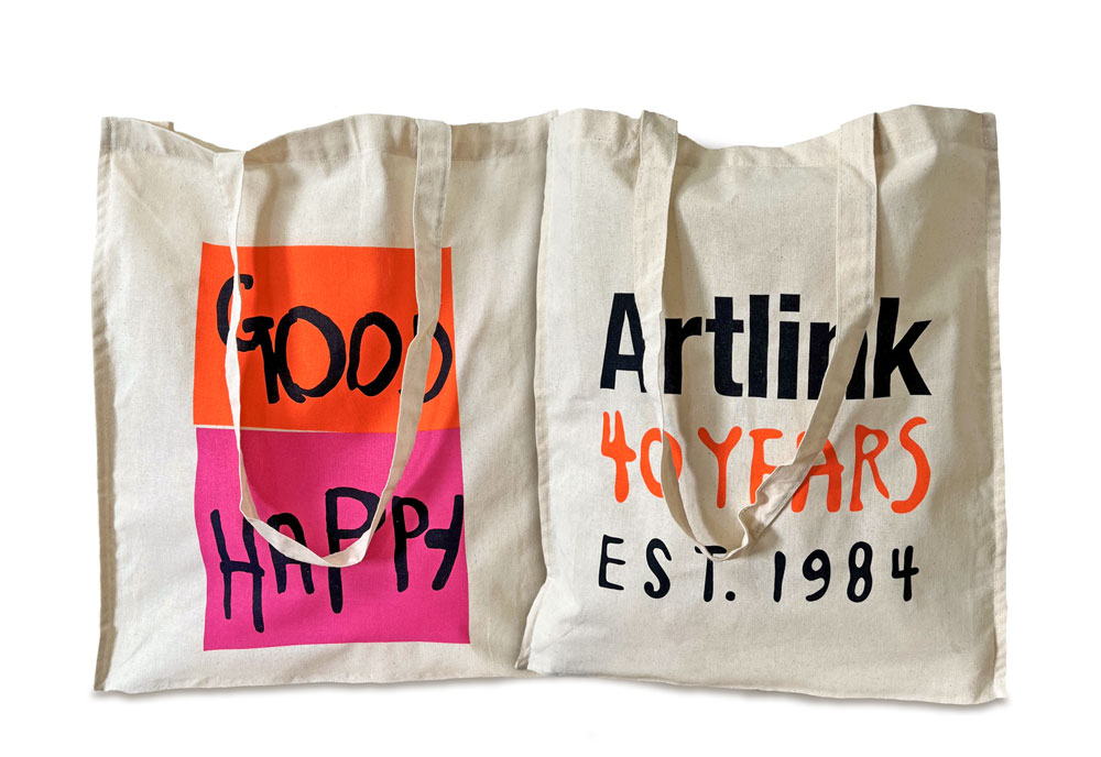 Beige cotton tote bag with long handles, printed on both sides with colourful design. One side has bright orange and pink rectangles with black text. The orange rectangle says 'Good' and the