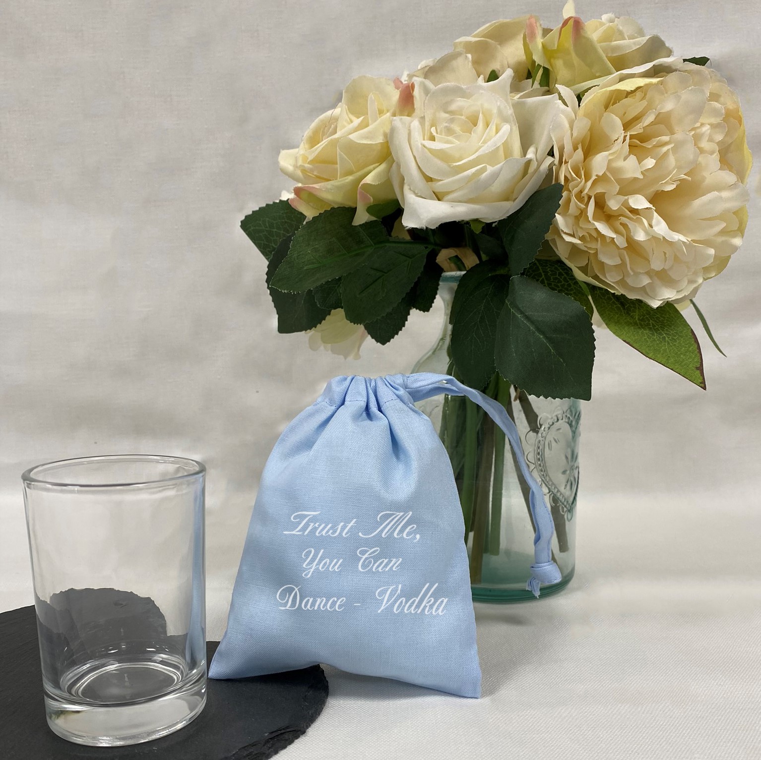 A picture containing a shot glass, a mini drawstring gift bag next to a bouquet of flowers