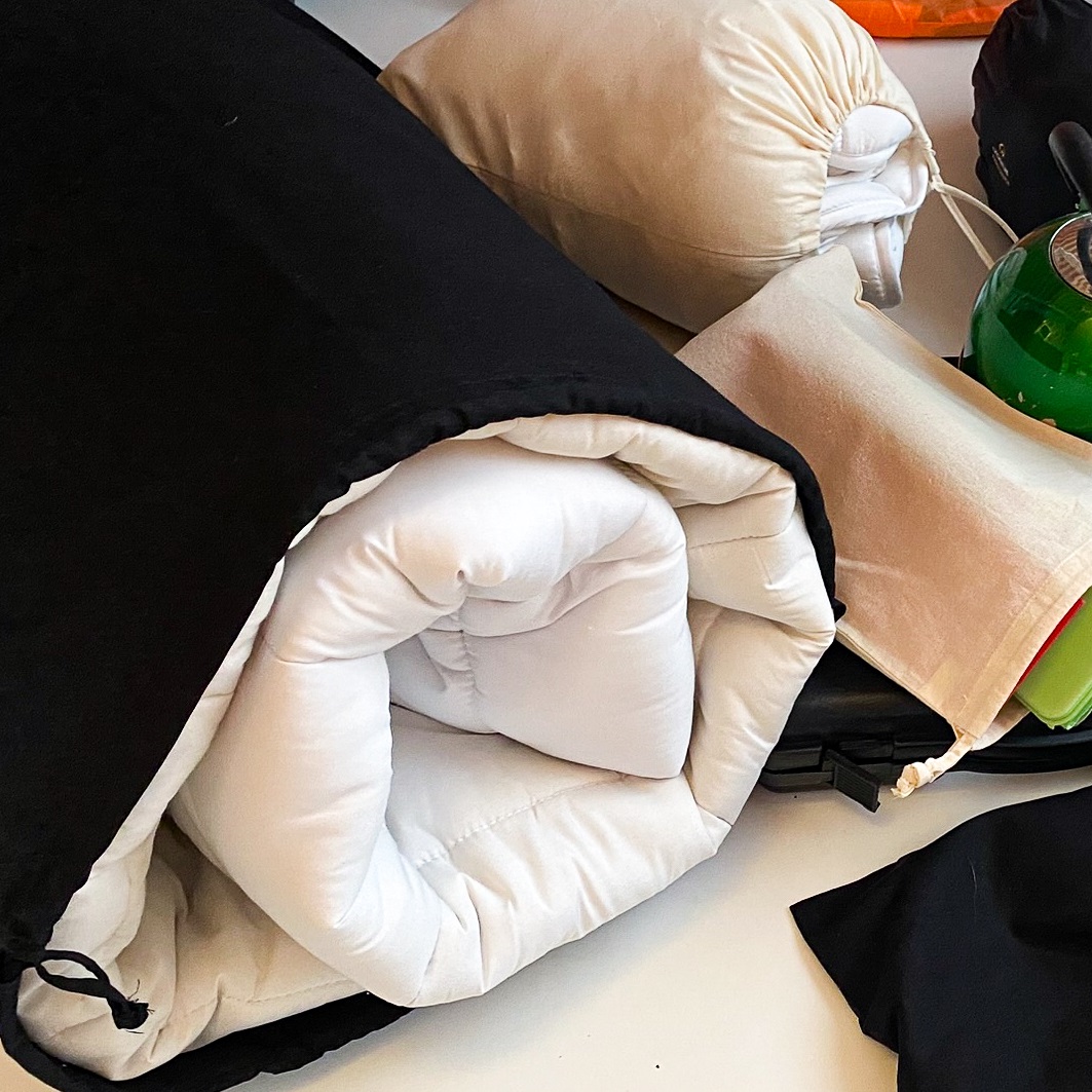 Cotton drawstring bags in a variety of sizes and colours being used to store different items: a large cotton sack holds a duvet while a smaller bag behind holds sheets.